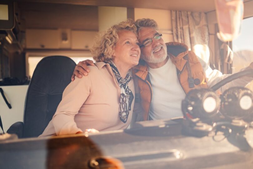 A senior man grips the steering wheel of a camper van while embracing a senior woman in the passenger seat.