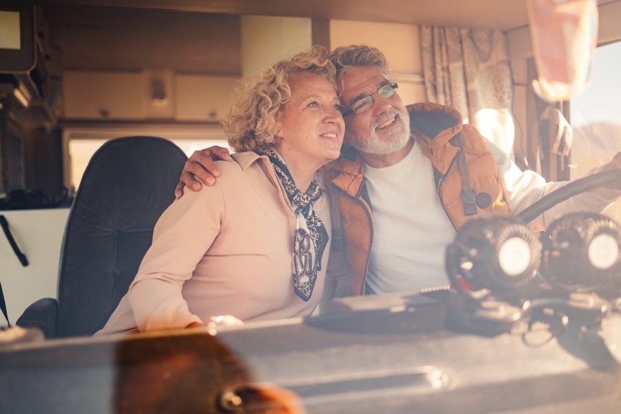A senior man grips the steering wheel of a camper van while embracing a senior woman in the passenger seat.