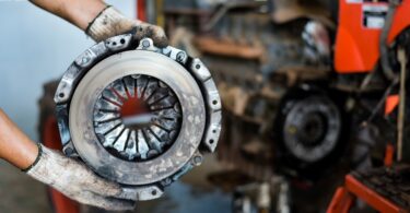 A close-up of a mechanic with white gloves holding up a used and dirty car clutch disc in front of a blurred vehicle.