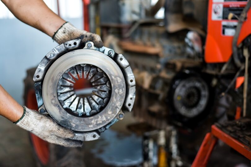 A close-up of a mechanic with white gloves holding up a used and dirty car clutch disc in front of a blurred vehicle.