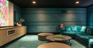 A modern teal velvet sofa and brown wooden circle tables in a home movie theater with a large TV on the wall.