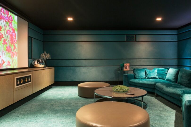 A modern teal velvet sofa and brown wooden circle tables in a home movie theater with a large TV on the wall.