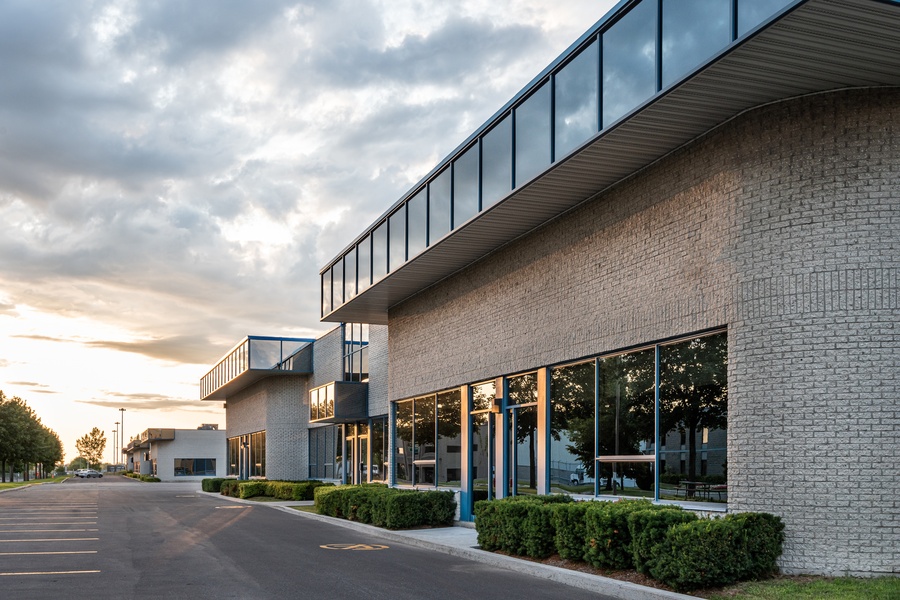 The exterior of a business contains brown bricks and large windows. A parking lot is in front of the building.