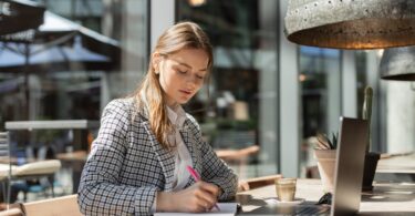 A stylish young businesswoman in a blazer focused on planning outside with coffee and a cactus nearby.