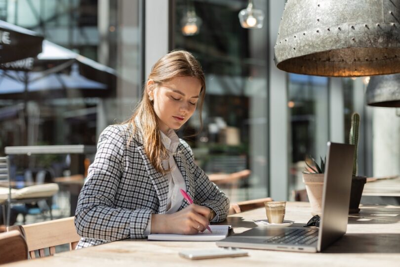 A stylish young businesswoman in a blazer focused on planning outside with coffee and a cactus nearby.