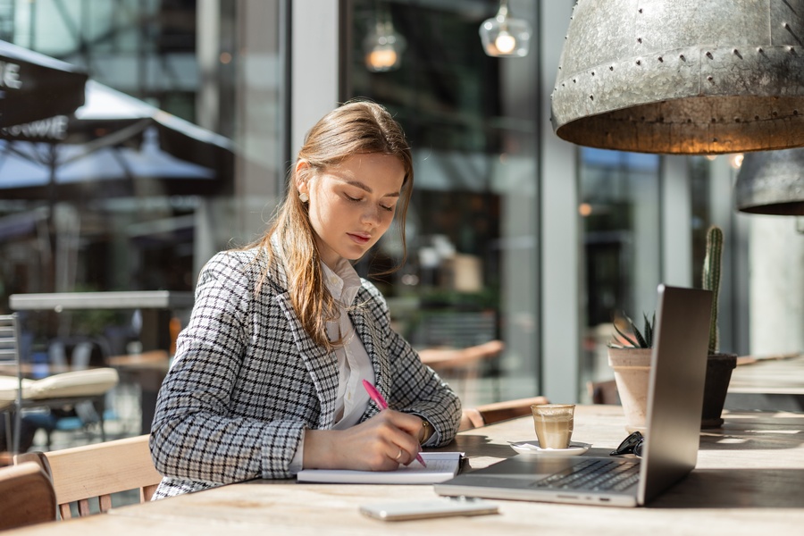 A stylish young businesswoman in a blazer focused on planning outside with coffee and a cactus nearby.