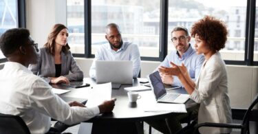 A diverse group of business professionals gathers in a meeting to discuss a business plan, each with their laptops.