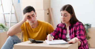 A stressed young couple assessing moving costs with a notebook and calculator. They're surrounded by cardboard boxes.