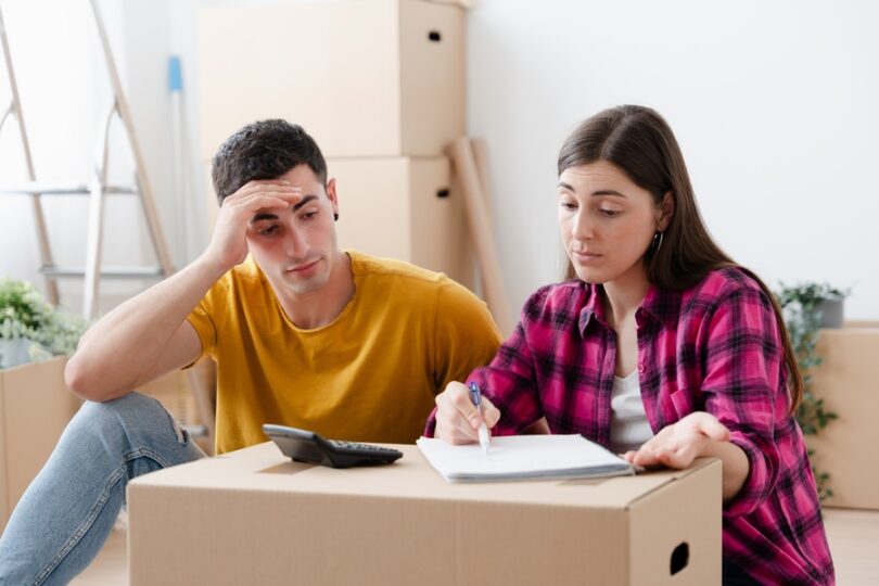 A stressed young couple assessing moving costs with a notebook and calculator. They're surrounded by cardboard boxes.