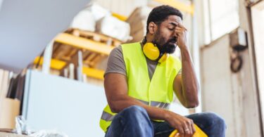 A tired industrial worker sitting with his head resting on his hand, holding his hard hat in the other hand.