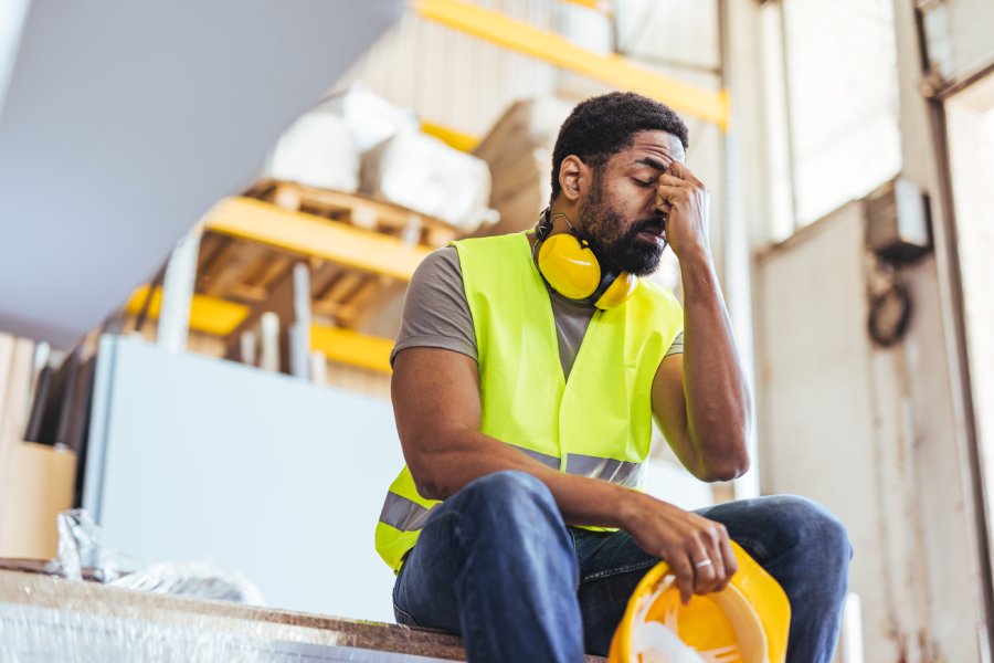 A tired industrial worker sitting with his head resting on his hand, holding his hard hat in the other hand.
