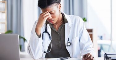 A stressed doctor in a lab coat sits at a desk in front of a laptop and notebook, resting her head against her right hand.