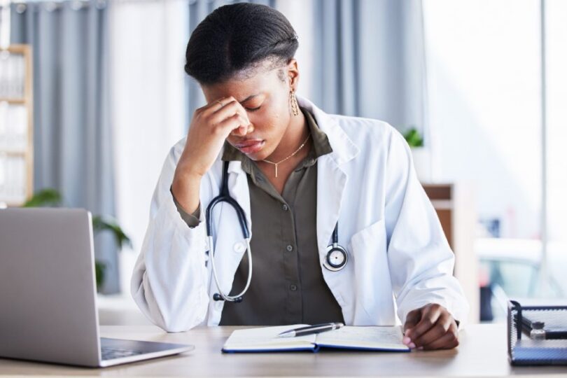 A stressed doctor in a lab coat sits at a desk in front of a laptop and notebook, resting her head against her right hand.