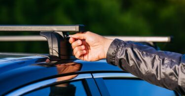 A close-up view shows a person wearing a black jacket using a key to lock the black roof rack on top of the vehicle.