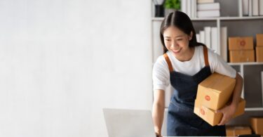 A young Asian businesswoman smiles while using a laptop, surrounded by boxes, and runs a successful online business.