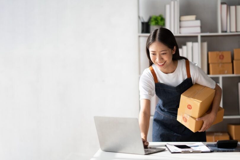A young Asian businesswoman smiles while using a laptop, surrounded by boxes, and runs a successful online business.
