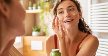 A close-up of a woman applying moisturizer on her face in her bathroom and smiling during her morning routine.