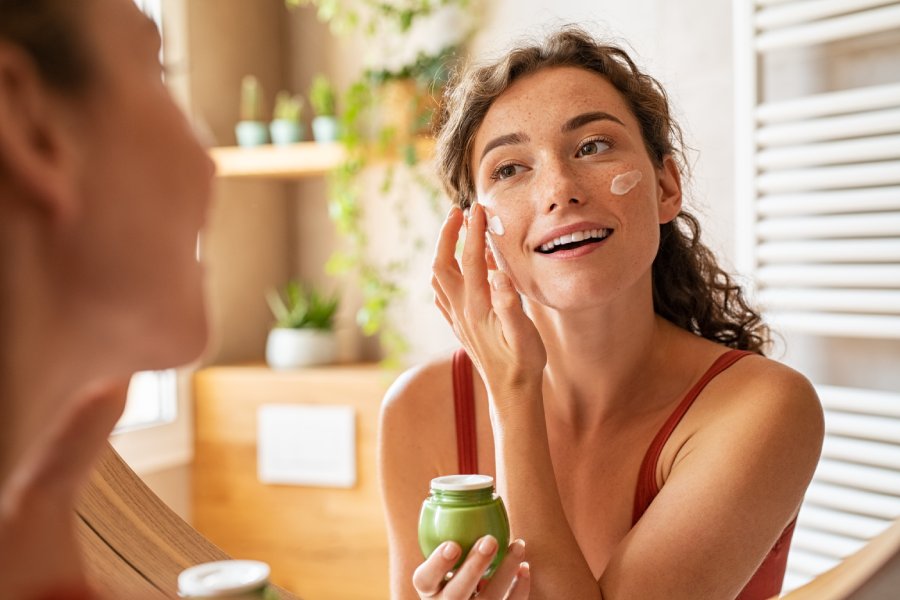 A close-up of a woman applying moisturizer on her face in her bathroom and smiling during her morning routine.