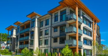 The exterior of a modern apartment building with balconies, wood accents, and landscaping on a sunny day.