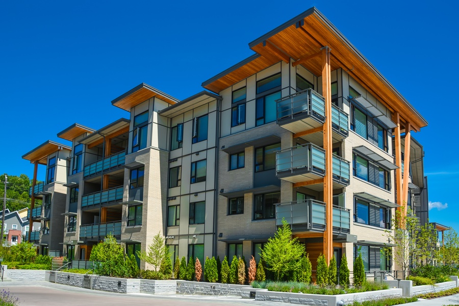 The exterior of a modern apartment building with balconies, wood accents, and landscaping on a sunny day.