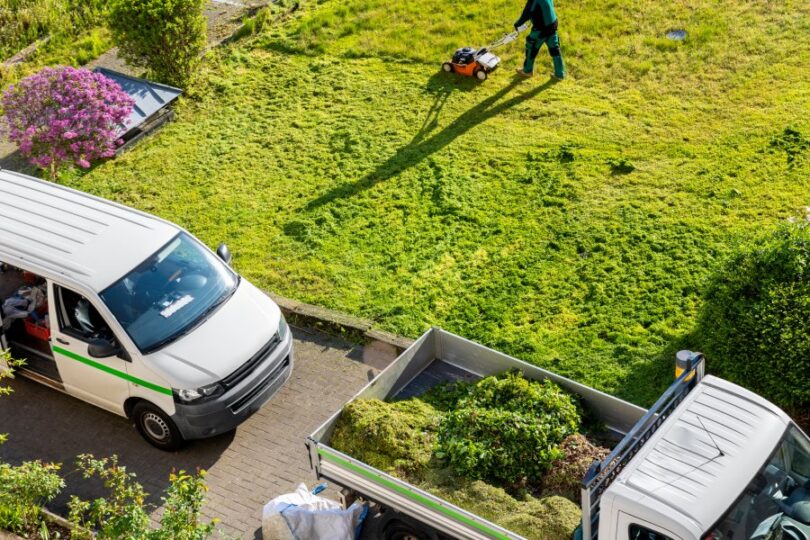 A professional landscaper mows the lawn of a business. There are two trucks parked near the lawn, one carries the brush.