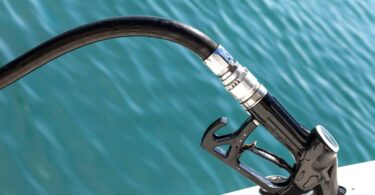 A close-up of a black fuel pump with the nozzle inside a boat. The water in the background is turquoise with small ripples.