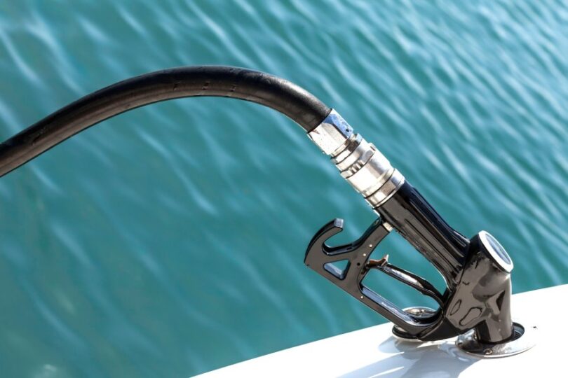 A close-up of a black fuel pump with the nozzle inside a boat. The water in the background is turquoise with small ripples.