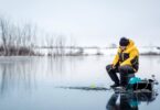 A person in thick outerwear sitting on a patch of ice on a lake and holding the end of a fishing rod to a hole in the ice.
