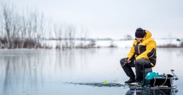 A person in thick outerwear sitting on a patch of ice on a lake and holding the end of a fishing rod to a hole in the ice.