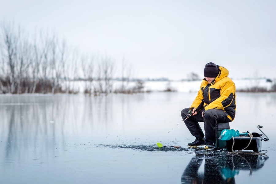 A person in thick outerwear sitting on a patch of ice on a lake and holding the end of a fishing rod to a hole in the ice.