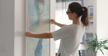 A woman with brown hair and glasses holds a long, tall painting of an abstract woman up to a wall in a home.