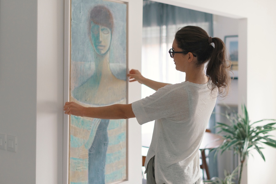A woman with brown hair and glasses holds a long, tall painting of an abstract woman up to a wall in a home.
