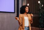 A woman in a white shirt, brown blazer, and jeans speaks into a microphone in front of a wall-mounted screen.