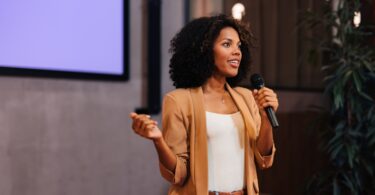 A woman in a white shirt, brown blazer, and jeans speaks into a microphone in front of a wall-mounted screen.