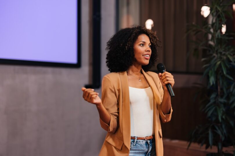 A woman in a white shirt, brown blazer, and jeans speaks into a microphone in front of a wall-mounted screen.