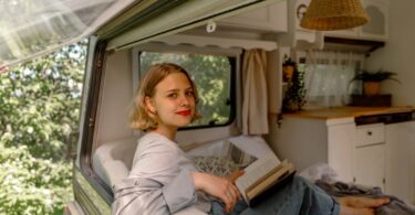 A woman sits in an RV with the window propped open. She has a book in her lap, and there are potted plants and curtains in the RV.