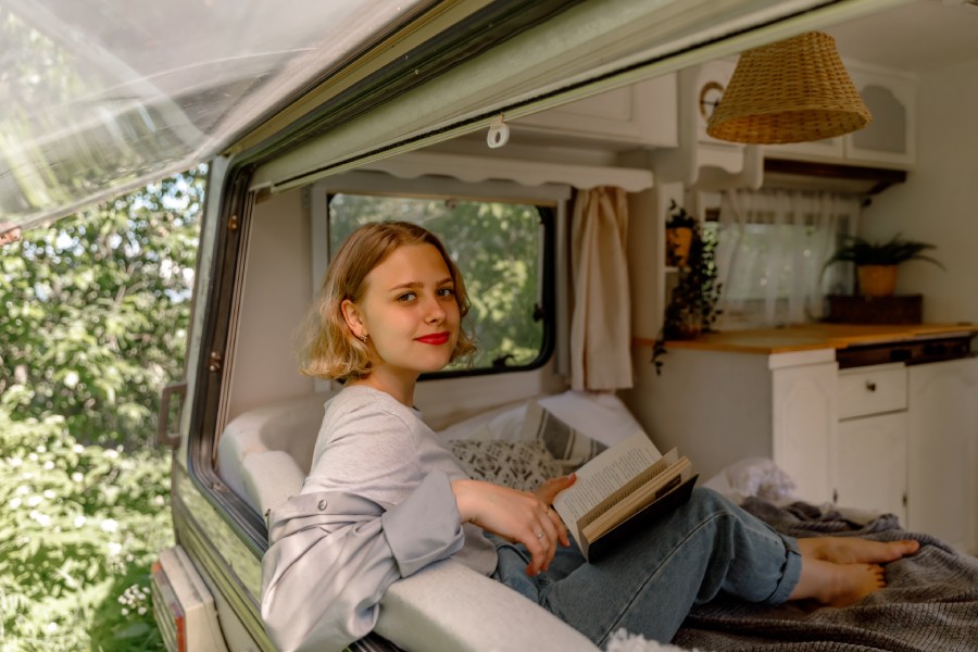 A woman sits in an RV with the window propped open. She has a book in her lap, and there are potted plants and curtains in the RV.