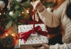 A gift recipient in a cream, cable-knit sweater holds a wrapped box as they undo the ribbon around the packaging.