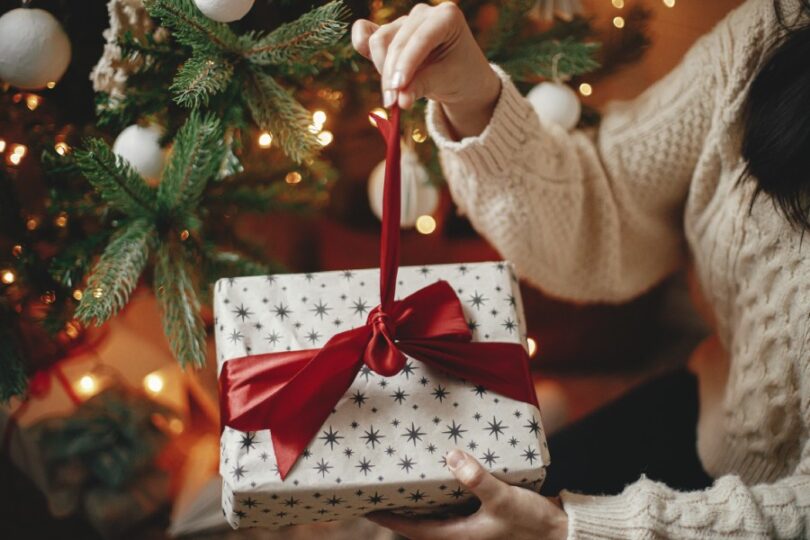 A gift recipient in a cream, cable-knit sweater holds a wrapped box as they undo the ribbon around the packaging.