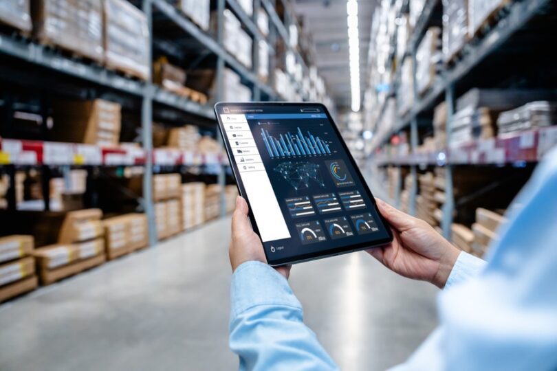 A businessman looks at a tablet in a large warehouse. The tablet has different business information on the screen.