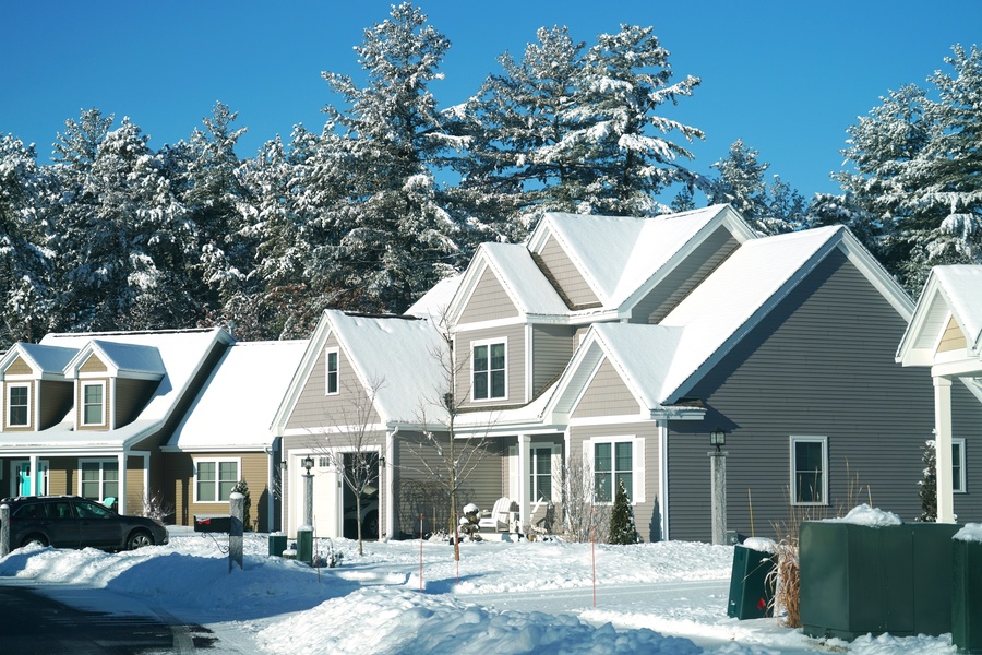 A row of houses covered in snow after a blizzard. There is snow on the lawn and on the trees behind the homes.