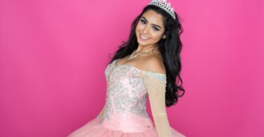 A smiling brown-haired teenager wears a strapless pink ballgown, paired with a statement necklace and a tiara.