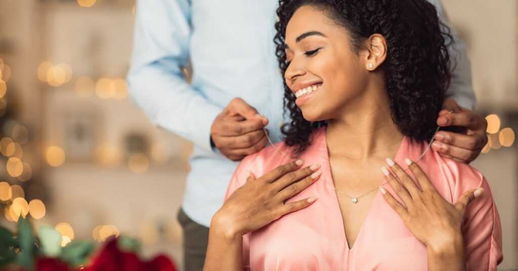 A person places a necklace on a smiling woman wearing a shiny pink blouse with blurred lights in the background.