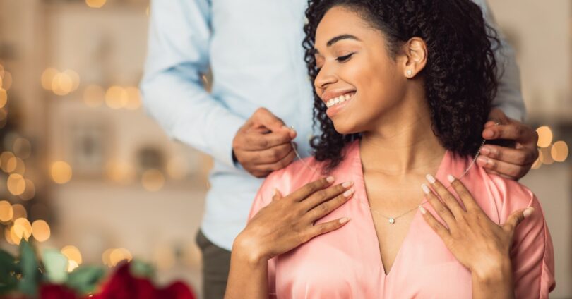 A person places a necklace on a smiling woman wearing a shiny pink blouse with blurred lights in the background.