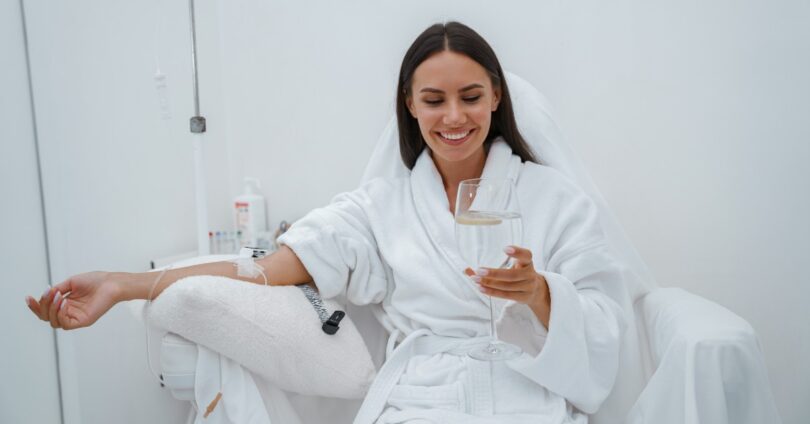 A young woman sits in a clinic chair as she scrolls her phone, smiling, receiving an IV therapy drip.