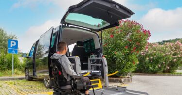 A man in a powered wheelchair sits in front of a black wheelchair-accessible van. The lift from the trunk is deployed.