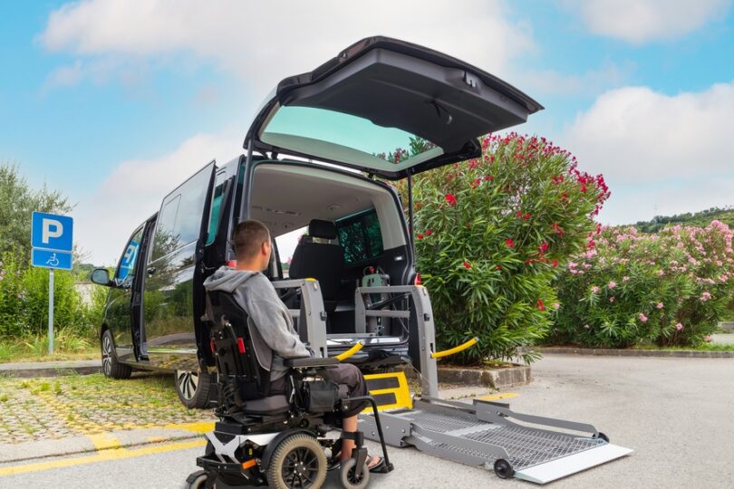 A man in a powered wheelchair sits in front of a black wheelchair-accessible van. The lift from the trunk is deployed.