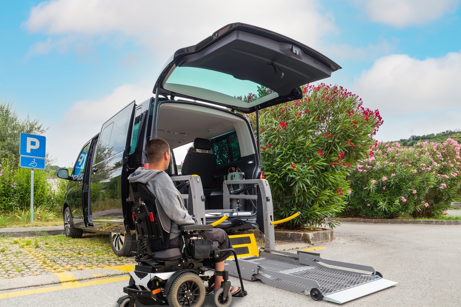 A man in a powered wheelchair sits in front of a black wheelchair-accessible van. The lift from the trunk is deployed.