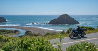 A motorcycle rider cruising along the California PCH next to the ocean. It's a sunny day outside and the ocean is blue.