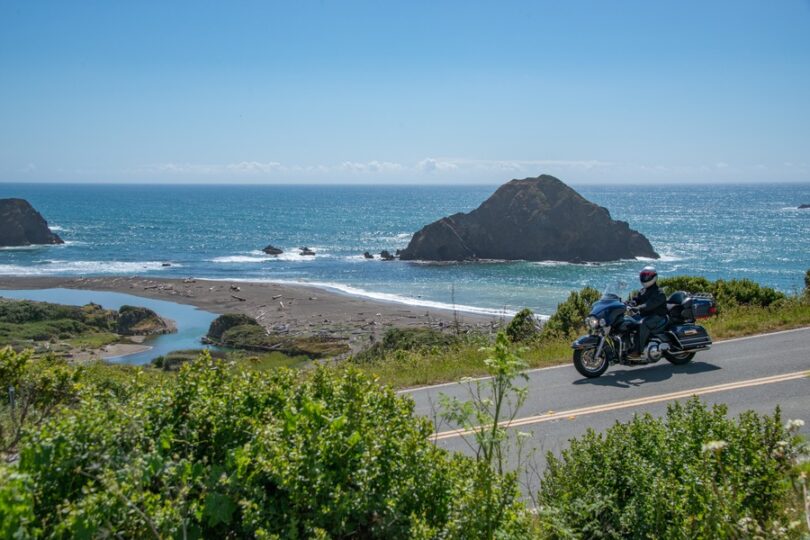 A motorcycle rider cruising along the California PCH next to the ocean. It's a sunny day outside and the ocean is blue.